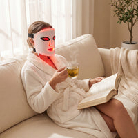 Relaxing in a sunlit living room, a person in a white robe sits on the sofa, wearing the LumiLift™ Red Light Therapy Silicone Face Mask, sipping tea and reading a book.