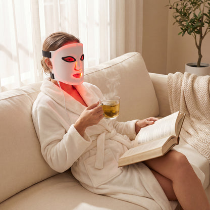Relaxing in a sunlit living room, a person in a white robe sits on the sofa, wearing the LumiLift™ Red Light Therapy Silicone Face Mask, sipping tea and reading a book.