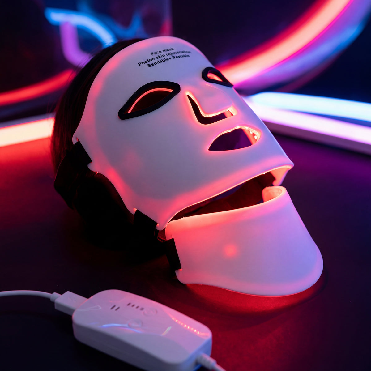 The LumiLift™ Red Light Therapy Silicone Face Mask features eye, nose, and mouth cutouts and emits red and purple lights in the dark. It connects to a control device via a white cord, with neon lights glowing in the background.