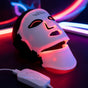 The LumiLift™ Red Light Therapy Silicone Face Mask features eye, nose, and mouth cutouts and emits red and purple lights in the dark. It connects to a control device via a white cord, with neon lights glowing in the background.