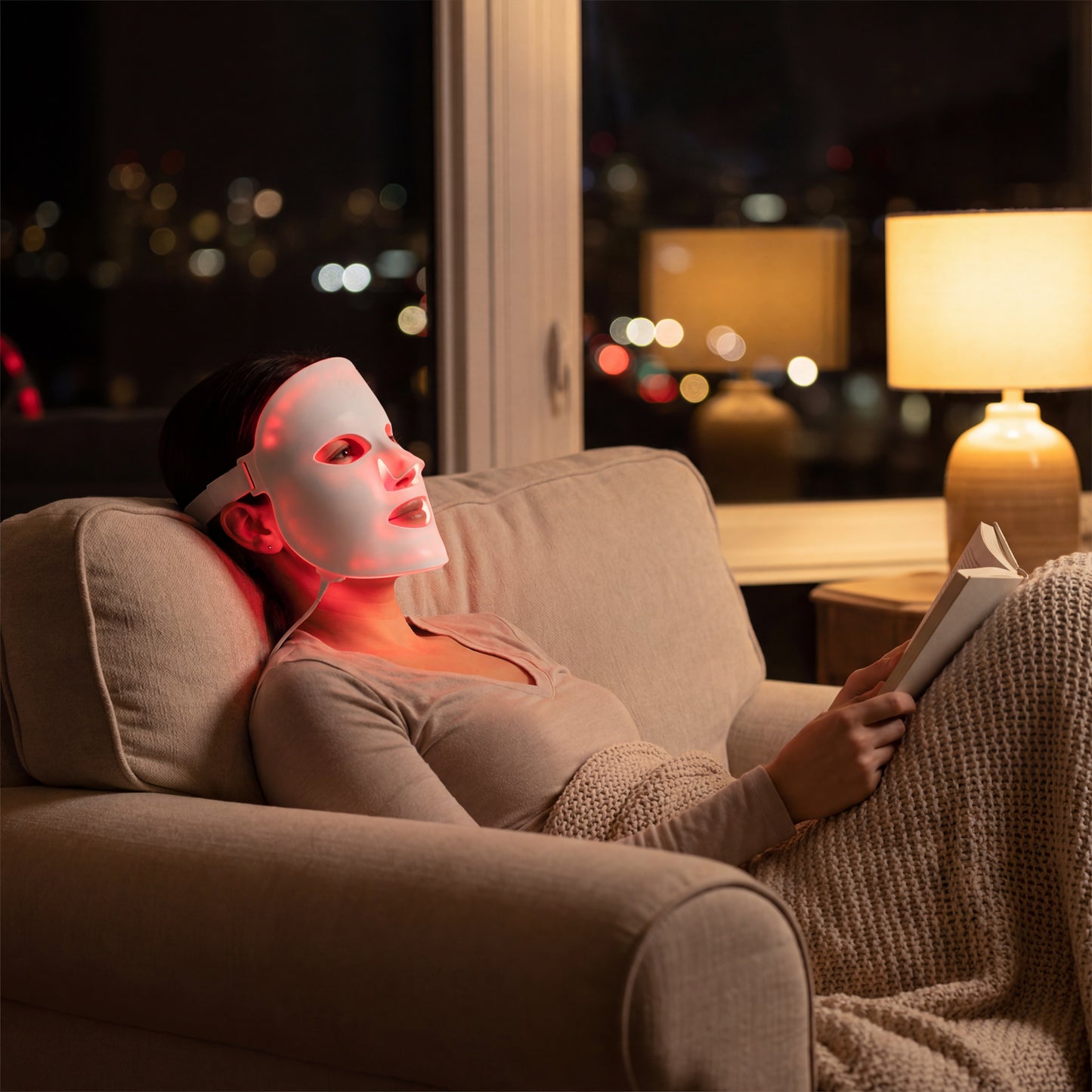 At night, a person sits on a couch with a blanket over their legs, wearing the illuminated LumiLift™ Red Light Therapy Silicone Face Mask and reading, as a soft lamp glows in the background.