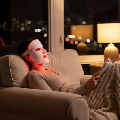 At night, a person sits on a couch with a blanket over their legs, wearing the illuminated LumiLift™ Red Light Therapy Silicone Face Mask and reading, as a soft lamp glows in the background.