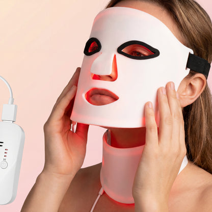 A woman wears the LumiLift™ Red Light Therapy Silicone Face Mask, secured with a strap and connected by a white cord to a control device, against a soft pink background.